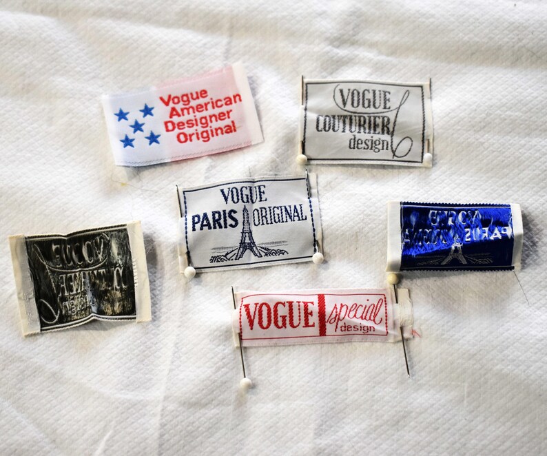 May include: A collection of vintage Vogue labels, each with unique text and designs. The labels feature phrases like "American Designer Original," "Paris Original," and "Special Design." The labels are pinned to a white textured surface.