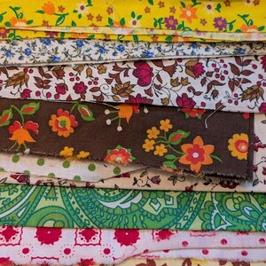 May include: A stack of colorful fabric swatches with floral and paisley patterns. The fabrics feature a variety of colors, including yellow, green, pink, and brown. Some fabrics have small polka dots. A ruler is visible at the bottom.