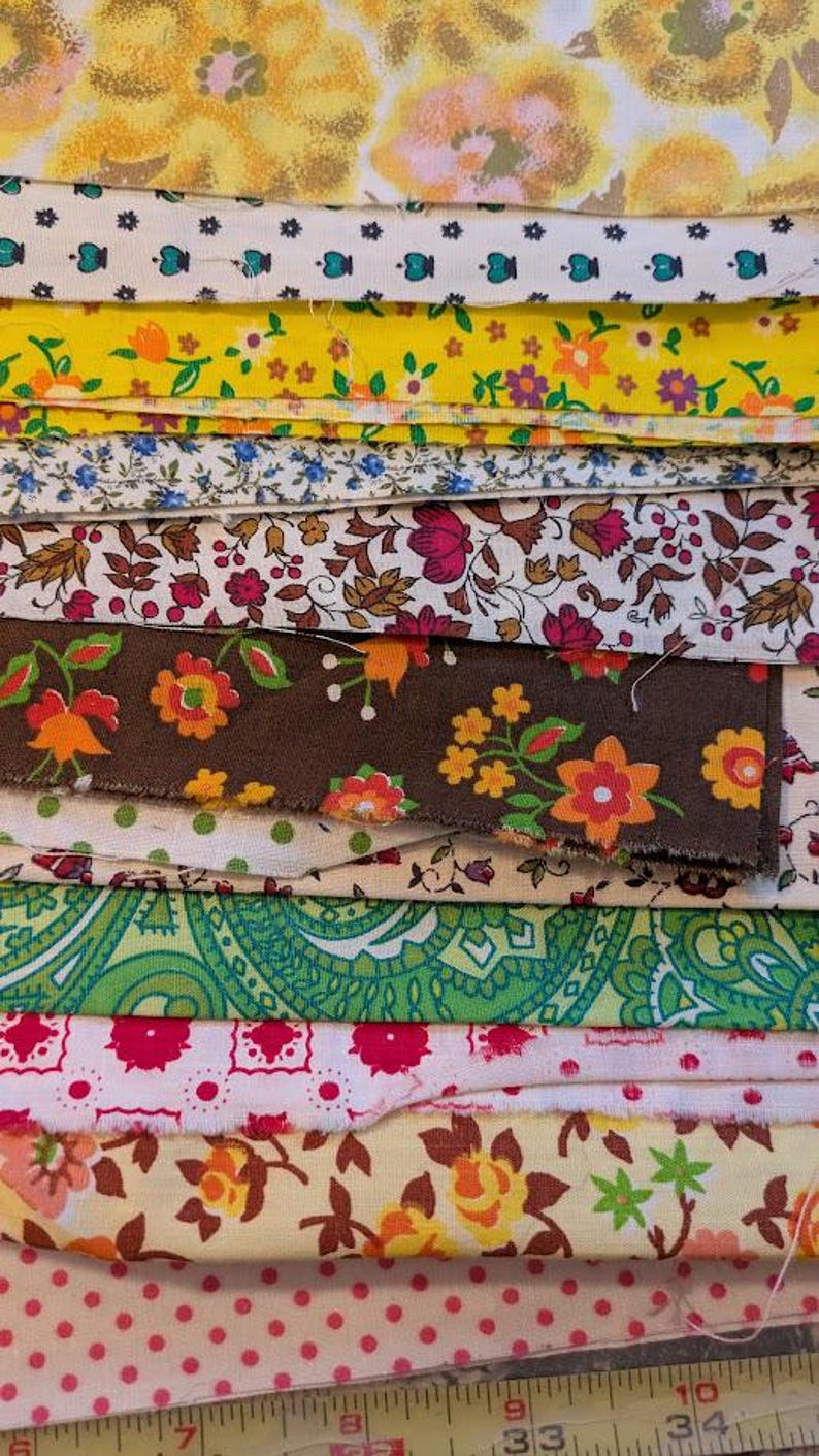 May include: A stack of colorful fabric swatches with floral and paisley patterns. The fabrics feature a variety of colors, including yellow, green, pink, and brown. Some fabrics have small polka dots. A ruler is visible at the bottom.