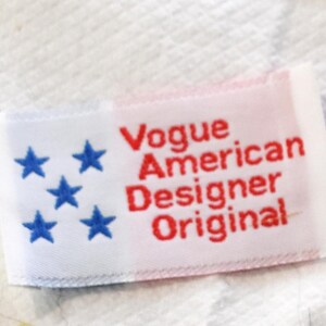 May include: A rectangular label with the text "Vogue American Designer Original" in red. The left side of the label has five blue stars on a white background, with a red background on the right.