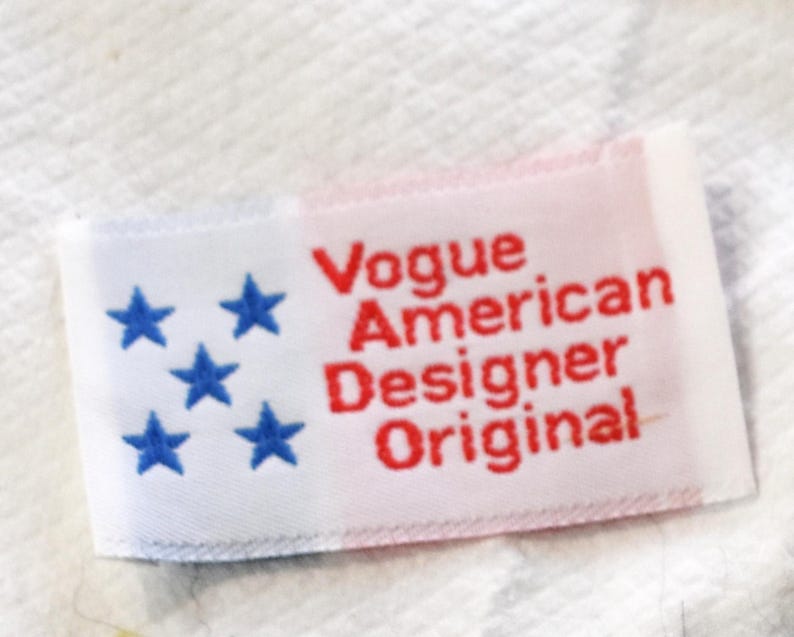 May include: A rectangular label with the text "Vogue American Designer Original" in red. The left side of the label has five blue stars on a white background, with a red background on the right.