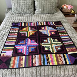 May include: A handmade log cabin quilt featuring four large central blocks with dark purple borders. The quilt is constructed from various colorful fabric strips in patterns including floral, striped, and solid designs, finished with a black binding.