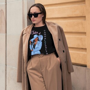 May include: A black t-shirt with a graphic of a woman wearing a blue suit and a hat. The text "A Sister of Gamma Phi Delta" is printed vertically on the right side of the shirt.