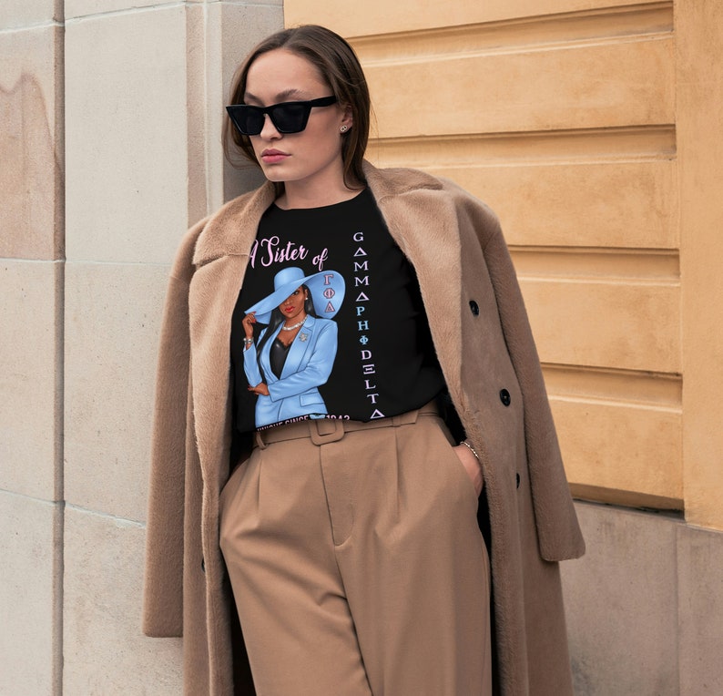 May include: A black t-shirt with a graphic of a woman wearing a blue suit and a hat. The text "A Sister of Gamma Phi Delta" is printed vertically on the right side of the shirt.