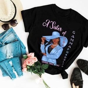 May include: Black t-shirt with a pink and blue illustration of a woman wearing a blue hat and suit. The text "A Sister of Gamma Phi Delta Unique Since 1943" is printed on the shirt.