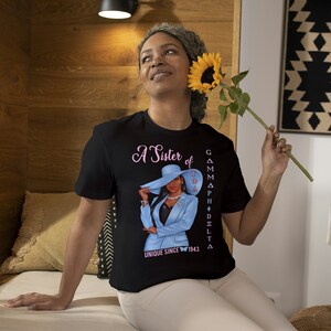May include: A black t-shirt with a pink and blue graphic of a woman wearing a blue suit and a hat. The text on the shirt reads "A Sister of Gamma Phi Delta Unique Since 1943".