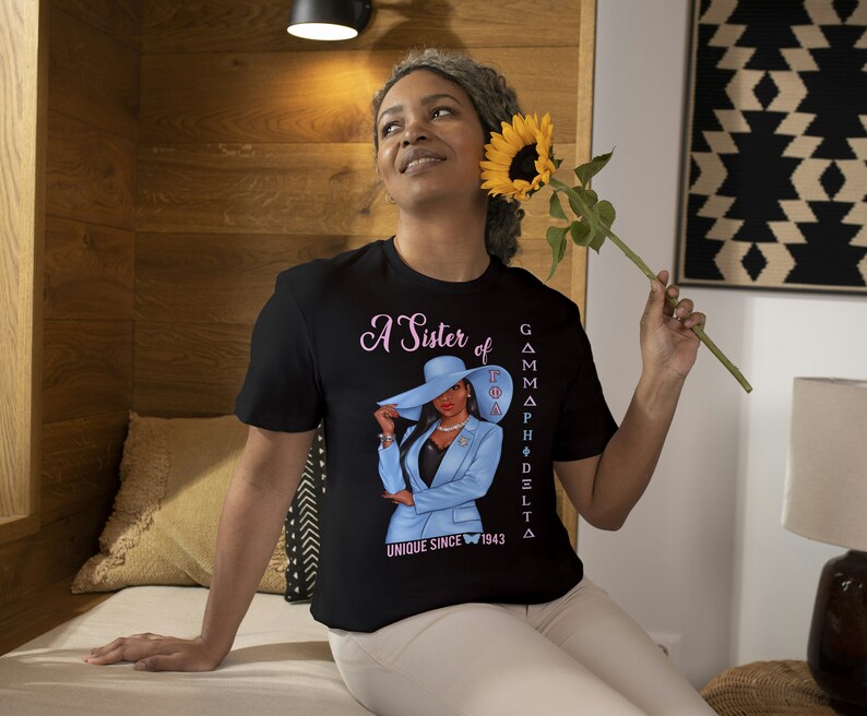 May include: A black t-shirt with a pink and blue graphic of a woman wearing a blue suit and a hat. The text on the shirt reads "A Sister of Gamma Phi Delta Unique Since 1943".