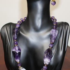 Op de afbeelding: Een amethisten ketting en bijpassende oorbellen, tentoongesteld op een zwarte mannequin. De ketting heeft grote, onregelmatig gevormde amethiststenen en kleinere, ronde kralen. Goudkleurige accenten voegen een vleugje elegantie toe. De oorbellen weerspiegelen het ontwerp van de ketting.