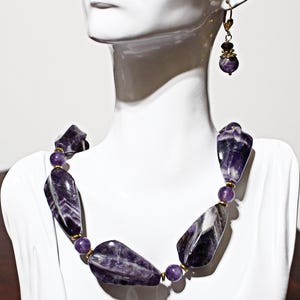 Op de afbeelding: Een ketting- en oorbellenset met grote, gepolijste amethiststenen in paarse en witte tinten. De ketting heeft grote, onregelmatig gevormde stenen, gescheiden door kleinere, ronde paarse kralen. De oorbellen hebben een vergelijkbaar ontwerp.