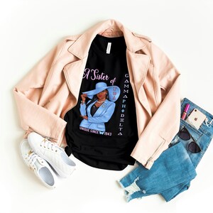 May include: A black t-shirt with a graphic of a woman wearing a blue suit and a hat. The text on the shirt reads "A Sister of Gamma Phi Delta Unique Since 1943".