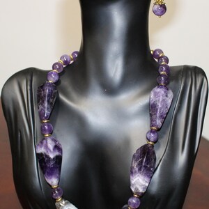 Op de afbeelding: Een amethisten ketting en bijpassende oorbellen, tentoongesteld op een zwarte mannequin. De ketting heeft grote, onregelmatig gevormde amethiststenen en kleinere, ronde kralen. Goudkleurige accenten voegen elegantie toe. De oorbellen spiegelen het ontwerp van de ketting.