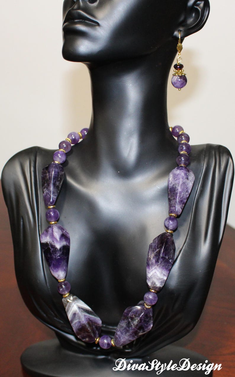 Op de afbeelding: Een amethisten ketting en bijpassende oorbellen, tentoongesteld op een zwarte mannequin. De ketting heeft grote, onregelmatig gevormde amethiststenen en kleinere, ronde kralen. Goudkleurige accenten voegen elegantie toe. De oorbellen spiegelen het ontwerp van de ketting.