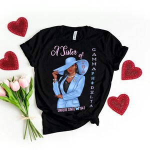 May include: Black t-shirt with a graphic of a woman wearing a blue suit and a hat. The text "A Sister of Gamma Phi Delta" and "Unique Since 1943" is printed on the shirt.