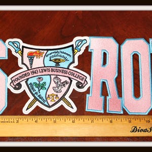 May include: Pink and light blue embroidered patches spelling out "SROR" with a crest. The crest features a shield with various symbols and the text "Founded 1943 Lewis Business College". A wooden ruler is placed below the patches.