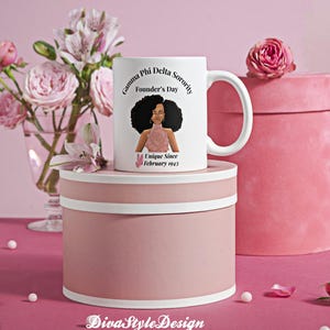 Peut inclure: Une tasse en céramique blanche avec un graphisme d'une femme avec un grand afro et le texte "Gamma Phi Delta Sorority Founder's Day Unique Since February 1943." La tasse est sur une boîte ronde rose avec un bord blanc, avec des fleurs roses en arrière-plan.