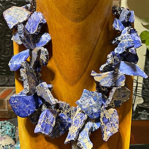 Puede incluir: Un collar de declaraci&oacute;n azul y dorado hecho de piedras grandes, de forma irregular y pulidas. El collar se muestra en un soporte de madera.