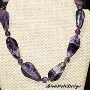 Op de afbeelding: Een ketting met grote, gepolijste amethiststenen in paarse en witte tinten, afgewisseld met kleinere ronde kralen. De ketting wordt getoond op een beige sieraden buste. De tekst "DivaStyleDesign" is zichtbaar.
