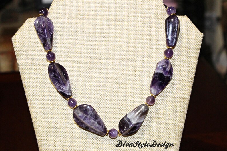 Op de afbeelding: Een ketting met grote, gepolijste amethiststenen in paarse en witte tinten, afgewisseld met kleinere ronde kralen. De ketting wordt getoond op een beige sieraden buste. De tekst "DivaStyleDesign" is zichtbaar.