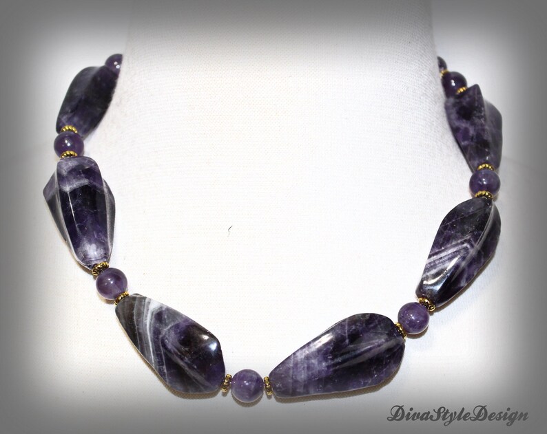 Op de afbeelding: Een ketting met grote, gepolijste amethiststenen in paarse en witte tinten. De stenen zijn onregelmatig van vorm en afgewisseld met kleinere, ronde paarse kralen en goudkleurige accenten. De ketting wordt gepresenteerd tegen een witte achtergrond.