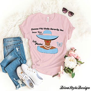 May include: A light pink t-shirt featuring a graphic of a woman wearing a blue hat and fur, with the text "Gamma Phi Delta Sorority, Inc. Unique Since 1943 Life Member." The shirt is paired with blue jeans and white sneakers.