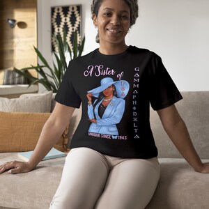 May include: A black t-shirt with a graphic of a woman wearing a blue suit and a hat. The text "A Sister of Gamma Phi Delta Unique Since 1943" is printed on the shirt.