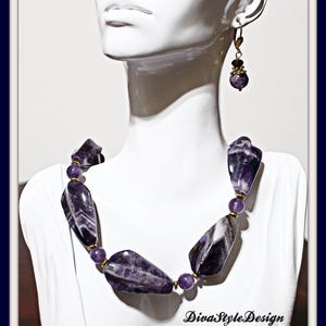 Op de afbeelding: Een ketting- en oorbellen set met gepolijste amethiststenen. De ketting heeft onregelmatig gevormde amethiststukken met goudkleurige accenten en kleinere paarse kralen. De oorbellen hebben een vergelijkbaar ontwerp met een bungelende amethist kraal. Het DivaStyleDesign logo is zichtbaar.