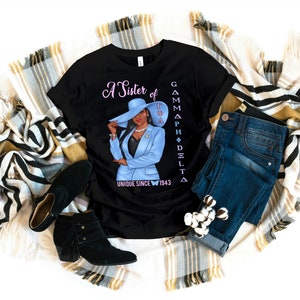 May include: Black t-shirt with a graphic of a woman wearing a blue suit and a hat. The text "A Sister of Gamma Phi Delta Unique Since 1943" is printed on the shirt.