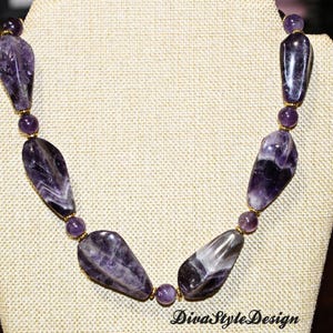 Op de afbeelding: Een ketting met grote, gepolijste amethiststenen in paarse en witte tinten, afgewisseld met kleinere ronde kralen. De ketting wordt getoond op een beige sieraden buste. De tekst "DivaStyleDesign" is zichtbaar.
