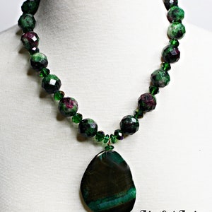 Puede incluir: Un collar de piedras preciosas verdes y rosas con un gran colgante de ágata verde. El collar está hecho de cuentas facetadas y está enhebrado en un alambre de cobre.