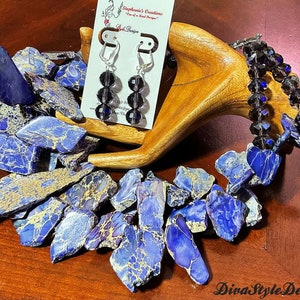 Puede incluir: Un collar de piedra marmoleada azul y dorada con un juego de pendientes a juego con cuentas de cristal negro. El collar est&aacute; hecho de piedras de forma irregular y los pendientes son de estilo colgante. La joyer&iacute;a se exhibe en un soporte de madera.
