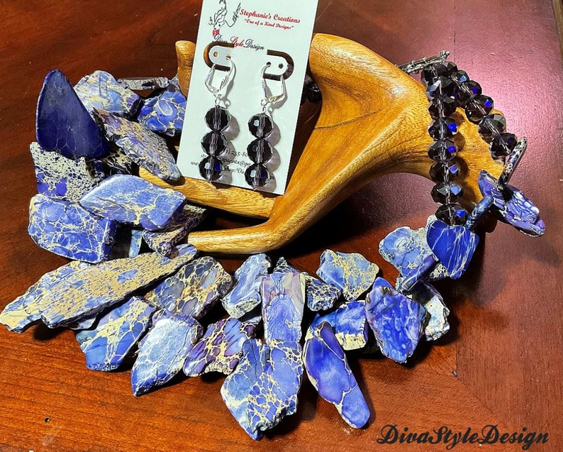 Puede incluir: Un collar de piedra marmoleada azul y dorada con un juego de pendientes a juego con cuentas de cristal negro. El collar est&aacute; hecho de piedras de forma irregular y los pendientes son de estilo colgante. La joyer&iacute;a se exhibe en un soporte de madera.