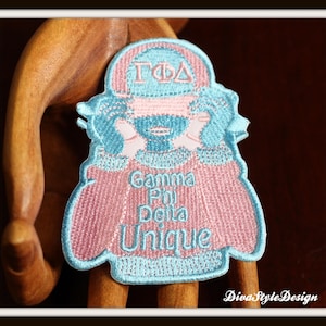 Może przedstawiać: Naszywka haftowana w kolorze niebieskim i różowym z postacią z kreskówki noszącą czapkę z napisem "GΦΔ". Postać ma na sobie różowy sweter z haftowanym napisem "Gamma Phi Delta Unique".