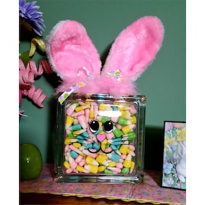 May include: A glass block vase with a bunny face design filled with pastel-colored candy corn. The vase has pink fuzzy bunny ears on top.