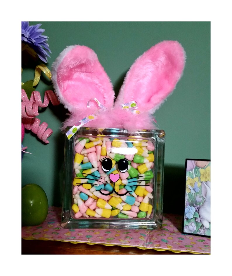 May include: A glass block vase with a bunny face design filled with pastel-colored candy corn. The vase has pink fuzzy bunny ears on top.