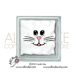 May include: White glass block with a black and pink cat face design. The cat has black eyes, a pink nose, and a black mustache.