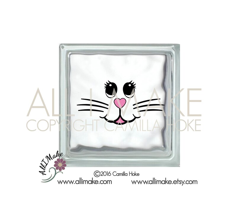 May include: White glass block with a black and pink cat face design. The cat has black eyes, a pink nose, and a black mustache.