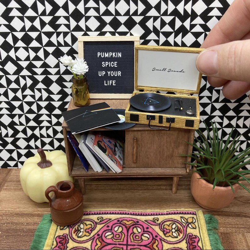 May include: A miniature record player with a black and gold design, a small pumpkin, a vase with white flowers, and a sign that reads "Pumpkin Spice Up Your Life".