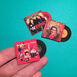 May include: Three miniature vinyl records with red and white Christmas album covers. The covers feature Elvis Presley, *NSYNC, and Perry Como. The records are black with a yellow label.