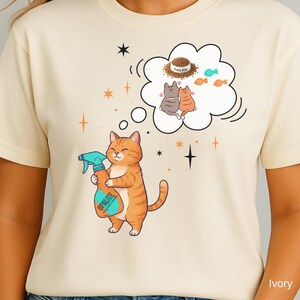 Könnte beinhalten: Elfenbeinfarbenes T-Shirt mit einer Cartoon-Katze, die eine Sprühflasche hält. Die Katze ist orange mit braunen Streifen. Eine Gedankenblase über der Katze zeigt zwei Katzen, Futter und Fische. Auf der Sprühflasche steht das Wort "PALITE".