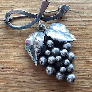 May include: A silver brooch in the shape of a grapevine with leaves and grapes. The brooch is made of metal and has a detailed design.