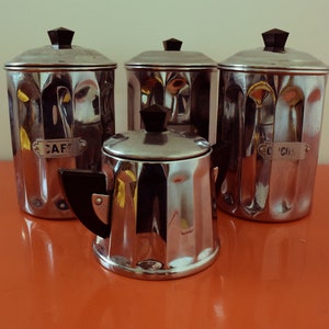 May include: Four silver canisters with black lids and labels. The labels read "CAF", "SUCRE", "THE", and "COCH".