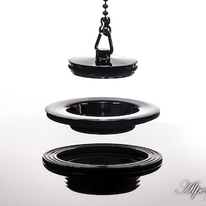 May include: A black sink drain stopper with a chain. The stopper is made of metal and has a round shape. The chain is attached to the stopper and is used to lift the stopper out of the drain.