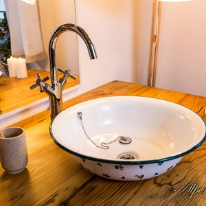 May include: A white porcelain sink with a green rim and floral pattern sits on a wooden countertop. A chrome faucet with a cross-shaped handle is mounted to the countertop. Two white candles are on the countertop to the right of the sink.