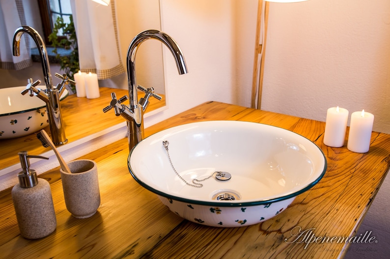 May include: A white porcelain sink with a green rim and floral pattern sits on a wooden countertop. A chrome faucet with a cross-shaped handle is mounted to the countertop. Two white candles are on the countertop to the right of the sink.