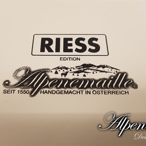 May include: A white enamelware pot with black lettering that reads "Riess Edition Alpenmaille Seit 1550 Handgemacht in &Ouml;sterreich Alpenmaille Design trifft Tradition".