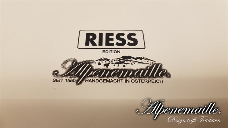 May include: A white enamelware pot with black lettering that reads "Riess Edition Alpenmaille Seit 1550 Handgemacht in &Ouml;sterreich Alpenmaille Design trifft Tradition".