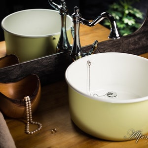 K&ouml;nnte beinhalten: Ein hellgelbes Keramik-Waschbecken mit wei&szlig;em Innenraum und einem verchromten Wasserhahn. Das Waschbecken steht auf einer Holztheke mit einer Holzschale und einer Perlenkette.