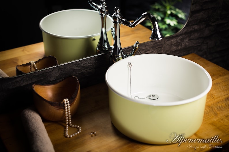 K&ouml;nnte beinhalten: Ein hellgelbes Keramik-Waschbecken mit wei&szlig;em Innenraum und einem verchromten Wasserhahn. Das Waschbecken steht auf einer Holztheke mit einer Holzschale und einer Perlenkette.
