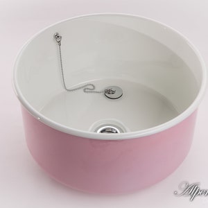 K&ouml;nnte beinhalten: Ein rosa Keramikwaschbecken mit wei&szlig;em Innenraum und einem verchromten Abflussstopfen mit Kette.