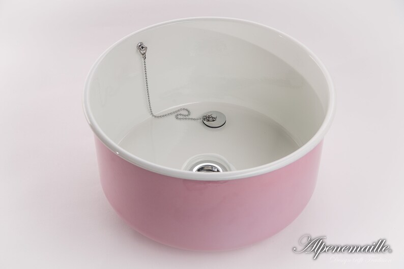 K&ouml;nnte beinhalten: Ein rosa Keramikwaschbecken mit wei&szlig;em Innenraum und einem verchromten Abflussstopfen mit Kette.
